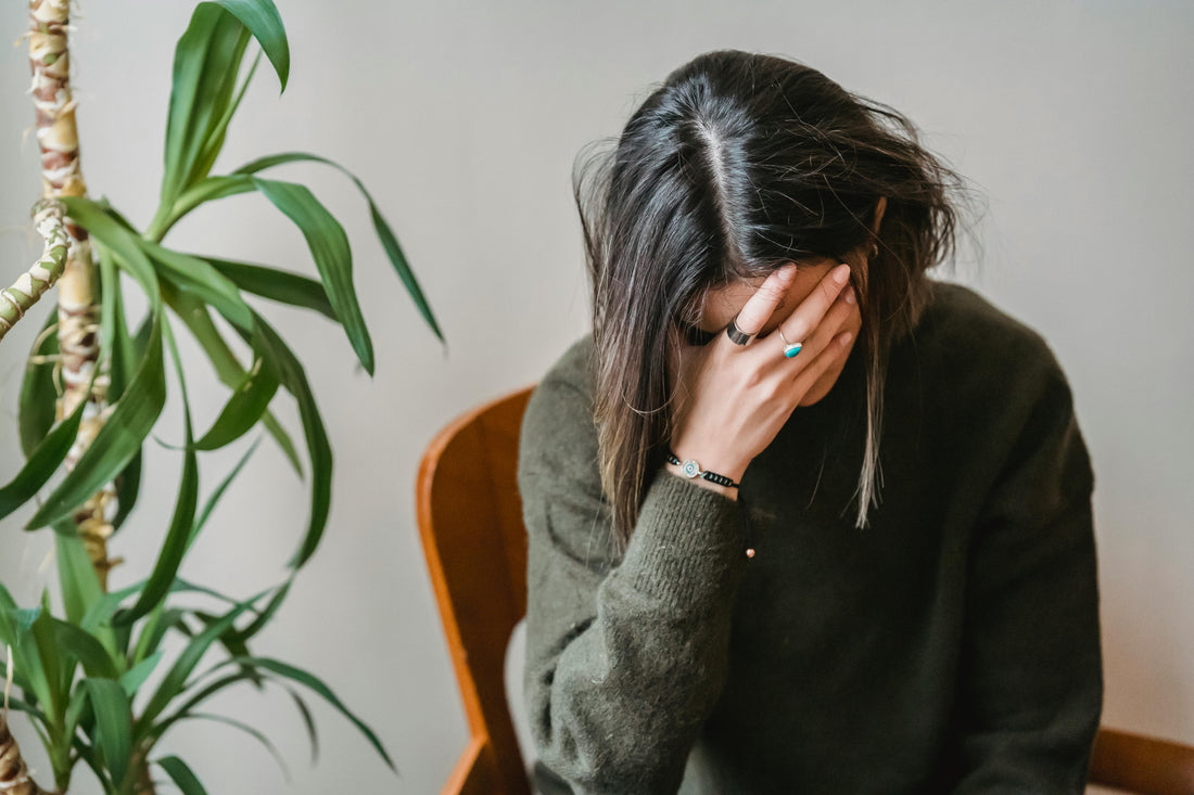 A single woman sitting on a chair next to a plant who is exhausted and is touching her forehead with her right palm because she has burnout from dating apps.
